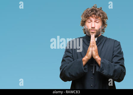 Handsome hispanic catholic priest man over isolated background praying with hands together asking for forgiveness smiling confident. Stock Photo