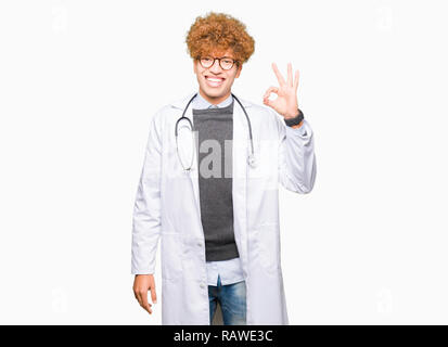 Young handsome doctor man wearing medical coat smiling positive doing ok sign with hand and fingers. Successful expression. Stock Photo