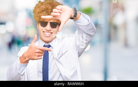 Young handsome scientist man wearing professional coat and sunglasses smiling making frame with hands and fingers with happy face. Creativity and phot Stock Photo