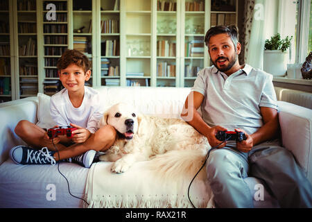 Father and son sitting on sofa with pet dog and playing video games