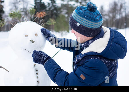 Young european man in warm clothes making snowman from snow outdoor. Having fun like a child at winter vacation. Stock Photo