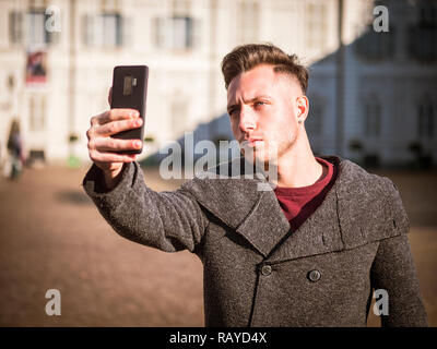 Half body shot of light brown haired, blue eyed young man wearing winter coat taking selfie photo with his smartphone in European city. Turin, Italy c Stock Photo