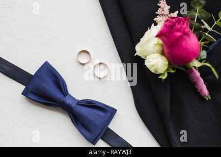 Close up top view of elegant modern formal male accessories. Blue bowtie, two golden wedding rings, flower boutonniere pinned to black jacket of suit. Stock Photo