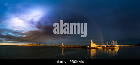 Howth Harbour in Dublin panorama under a dramatic stormy cloudy sky and rainbow spreading from the Eye of Ireland across to a lighthouse and sail boat Stock Photo