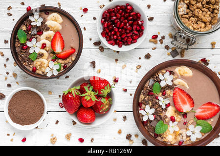chocolate banana protein smoothie bowls with granola, strawberry and pomegranate seeds decorated with flowers served for breakfast with spoons on whit Stock Photo