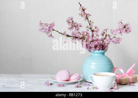 Pink cherry blossom flower bouquet with coffee cup, macaroons and gift box in blue vintage vase on white wooden background. holiday still life Stock Photo