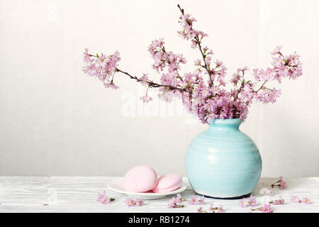 Pink cherry blossom flower bouquet with macarons in blue vintage vase on white wooden background. holiday still life Stock Photo