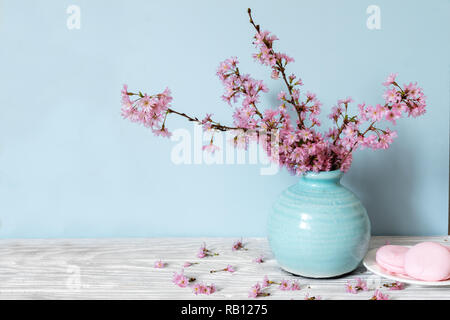 still life with bouquet of blooming cherry branches or sakura in a vase with macarons on blue background. holiday concept Stock Photo