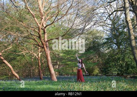 A fashionably dressed young lady with long brunette hair and red flares poses in a wood. The English woodland is carpeted with a sea of bluebells and  Stock Photo