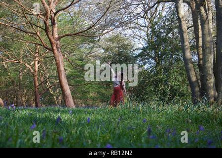 A fashionably dressed young lady with long brunette hair and red flares poses in a wood. The English woodland is carpeted with a sea of bluebells and  Stock Photo