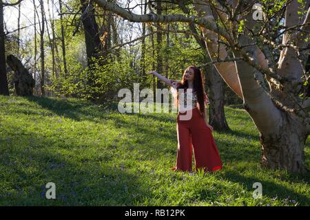 A fashionably dressed young lady with long brunette hair gestures, posing by a silver birch tree in an English woodland during a Spring evening Stock Photo
