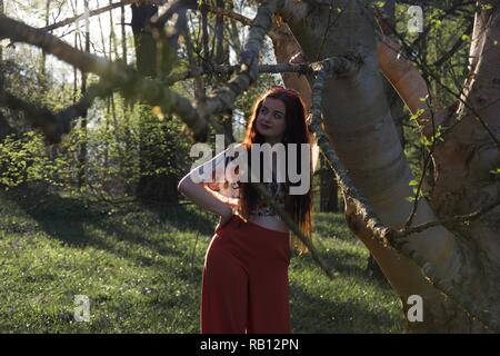 A young lady with long brunette hair and fashionably dressed in red flared trousers leans against a silver birch tree in an English woodland in spring Stock Photo