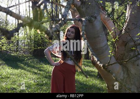 A young lady with long brunette hair and fashionably dressed in red flared trousers leans against a silver birch tree in an English woodland in spring Stock Photo