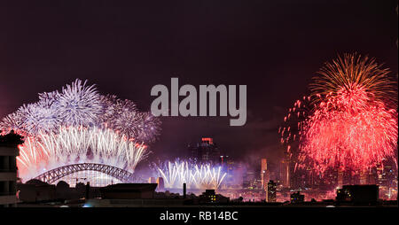 The top of the Sydney Harbour bridge with bright New Year light show fireworks against dark midnight sky next to Sydney city CBD towers and bright red Stock Photo