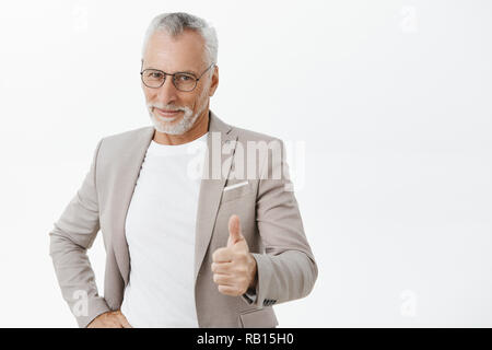 Nice job son. Portrait of proud and pleased good-looking old father in elegant suit and glasses showing thumbs up and smiling delighted from good positive result showing like gesture over grey wall Stock Photo