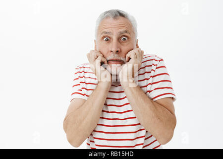 Portrait of intense anxious and scared charismatic senior man with grey beard and hair holding hands near mouth pursing lips and popping eyes at camera staring frightened and terrified at camera Stock Photo