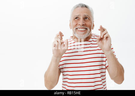 Optimistic happy and charismatic old male pensioner in striped t-shirt raising hands crossing fingers for good luck looking at upper left corner with hopeful smile making wish over white wall Stock Photo