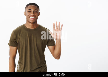 Friendly attractive nice african american male friend raising palm to wave smiling broadly saying hello, greeting friends in cafe welcoming or inviting people to party, posing over gray background Stock Photo
