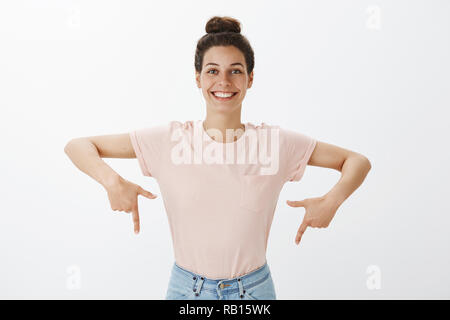 Waist-up shot of cheerful bright and happy charismatic beautiful european female with nice cheerful smile pointing down with raised fingers as showing proudly project she made over gray wall Stock Photo