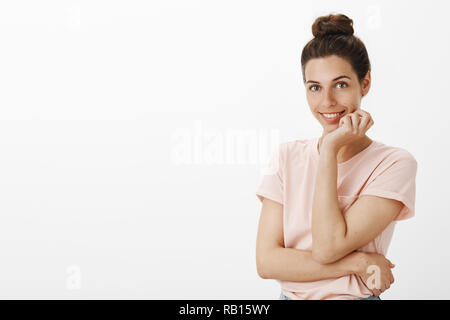 Attractive stylish and confident flirty european woman with cute hair bun in t-shirt smiling joyfully and tender at camera as holding hand on jawline posing and gazing at camera as seducing Stock Photo