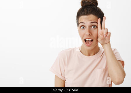 Hey chill and enjoy life. Carefree good-looking relaxed and charismatic stylish european woman in pink t-shirt raising eyebrows playfully as showing peace sign near cheek open mouth in yeah sound Stock Photo