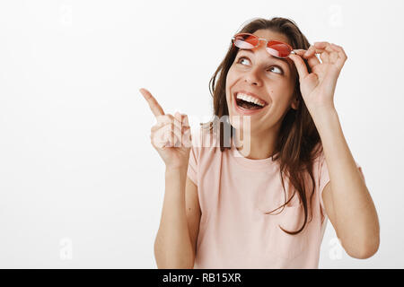 Amused girl looking at funny cat on tree loving animals taking off sunglasses as observing pointing at upper left corner smiling and laughing amazed and joyful, having fun against gray background Stock Photo