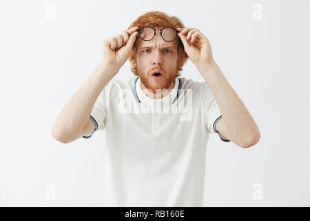 Guy getting closer look at confusing thing. Intense doubtful and questioned redhead intrigued guy with beard taking off glasses holding them on forehead folding lips and opening mouth from interest Stock Photo