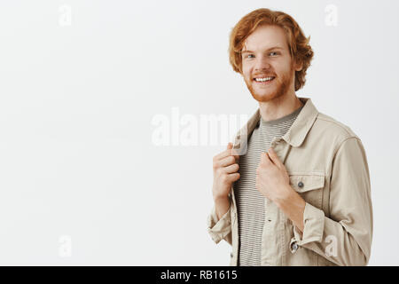 Man turning near mirror liking new outfit getting dressed for tonight date. Portrait of pleased happy redhead male model with beard trying on new clothes touching jacket and smiling broadly satisfied Stock Photo