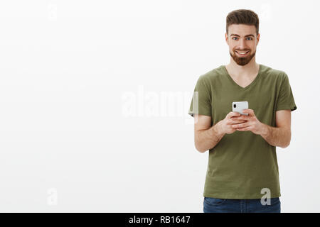 Devious and excited good-looking mature bearded male in olive t-shirt holding smartphone and looking pleased at camera grinning as having interesting idea of next post online over white wall Stock Photo