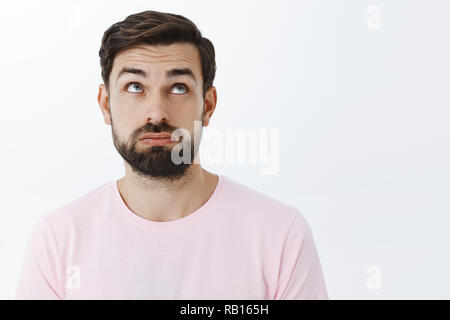 Close-up shot of unlucky, tired and upset attractive young bearded male with beard raising eyebrows in despair looking up with regret and disappointment, standing unconfident and exhausted Stock Photo
