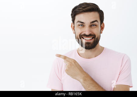 Amused joyful and pleased upbeat young man with beard and blue eyes laughing and smiling broadly as pointing left at cool place where he had fun, standing entertained and delighted over gray wall Stock Photo