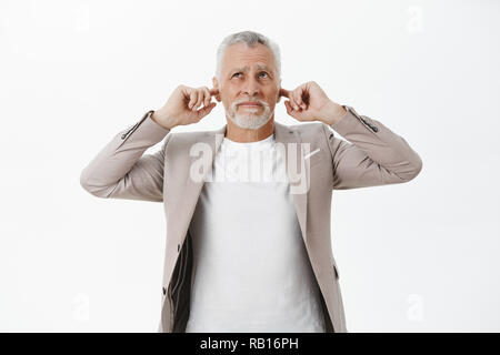 Portrait of old businessman being disturbed by loud noise comming upwards closing ears with index fingers like earplugs looking up worried and displeased wish sound or yell over white background Stock Photo