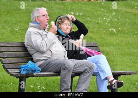Middle aged to elderly couple of people sitting on a wooden park bench wearing coats eating ice creams, on a cold day in Spring in the UK. Stock Photo