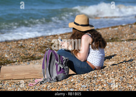 Young woman wearing sun hat sitting on a beach using a smartphone on a hot day in Summer in the UK. Stock Photo