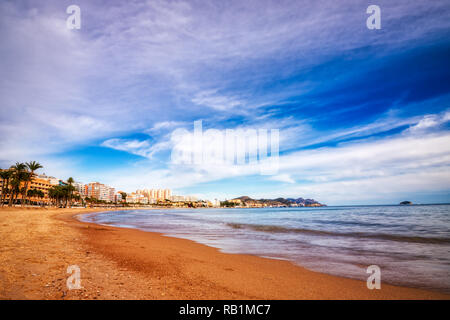 Low angle shot of the waves coming up on the snady beach of Villajoyosa Spain with a blue sky, white clouds and the buildings lining the shore in the  Stock Photo