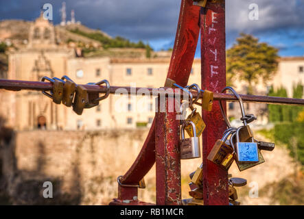 A group of love locks on a small part of a red bridge in Cuenca Spain with buildings, cliffs, trees, clouds and sky in soft focus in the distance (nam Stock Photo
