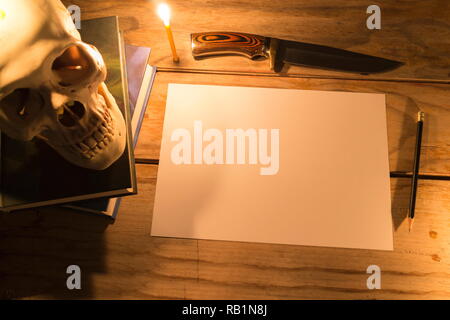 Human skull with candle light and white paper with pencil and knife on wooden table, Decorated for Halloween Theme with copy space. Stock Photo