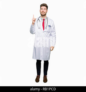 Young handsome doctor man wearing medical coat pointing finger up with successful idea. Exited and happy. Number one. Stock Photo