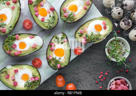 Keto diet dish: Avocado boats with ham cubes, quail eggs and cherry tomatoes on marble serving board on dark Stock Photo