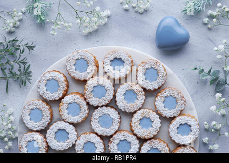 Top view of flower Linzer cookies with blue glazing on light blue background decorated with white flowers and ceramic heart. Birthday or Valentine bac Stock Photo