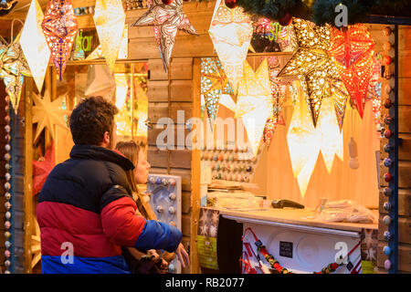People at Christmas Market stall (potential customers) looking at paper star lights (lanterns) on display casting warm golden glow - York, England, UK Stock Photo