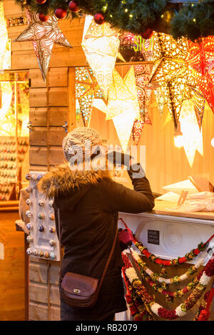 Woman at Christmas Market stall (potential customer) looking at paper star lights (lanterns) on display, casting warm golden glow - York, England, UK. Stock Photo