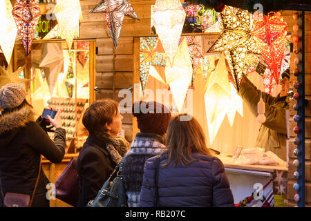 People at Christmas Market stall (potential customers) looking at paper star lights (lanterns) on display casting warm golden glow - York, England, UK Stock Photo
