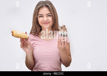 Young and cheerful woman hold yellow eclair and chocolate cake in hands. She look on camera. Isolated on grey background. Stock Photo
