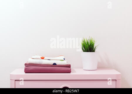 On a pink dresser there is a stack of bed linen and a home plant stands. Stock Photo