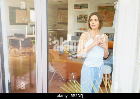 Portrait of young pretty woman wearing pajamas while standing with a cup of coffee behind window and dreaming. Stock Photo