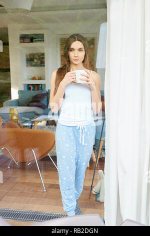 Portrait of young pretty woman wearing pajamas while standing with a cup of coffee near window and dreaming. Stock Photo