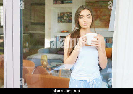 Portrait of young pretty woman wearing pajamas while standing with a cup of coffee  behind window and dreaming. Stock Photo