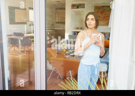 Portrait of young pretty woman wearing pajamas while standing with a cup of coffee behind window and dreaming. Stock Photo