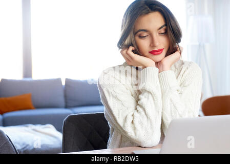 Portrait of smiling young woman sitting behind her notebook and looking thoughtfully while studying online. Stock Photo